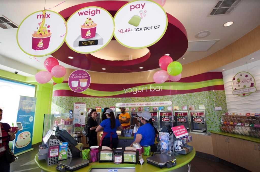 Menchie’s workers behind the circular counter help talk with customers against a backdrop of the curved wall of frozen yogurt dispensers.
