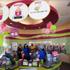 Menchie’s workers behind the circular counter help talk with customers against a backdrop of the curved wall of frozen yogurt dispensers.