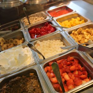 A cafeteria-style serving bar shows bins of toppings including fruit, cake bites and whipped cream.