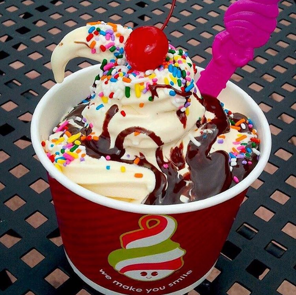 A cup of frozen yogurt with rainbow sprinkles, chocolate sauce and a cherry on top.