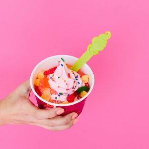 A cup of light pink frozen yogurt is topped with rainbow sprinkles, mochi and gummy worms, set against a pink background.
