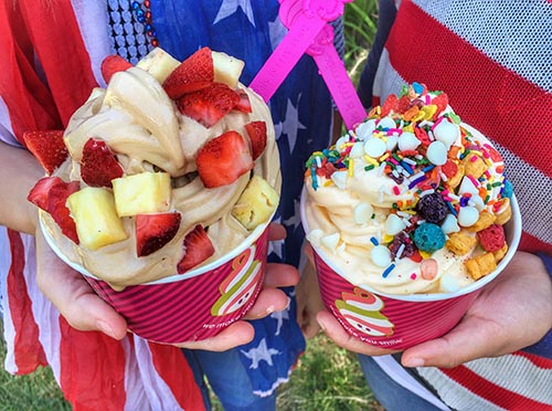 Two children hold Menchie’s frozen yogurt cups, one topped with strawberry and pineapple and the other topped with white chocolate chips, cereal and sprinkles.